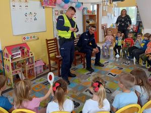Policjant założył kamizelkę odblaskową na mundur, dzieci siedzą na krzesełkach i słuchają pogadanki. Spotkanie odbywa się w sali szkolnej